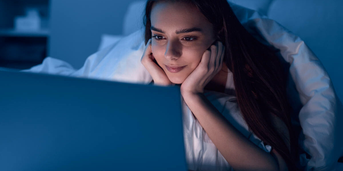 Young Hispanic lady watching video on laptop while resting on bed